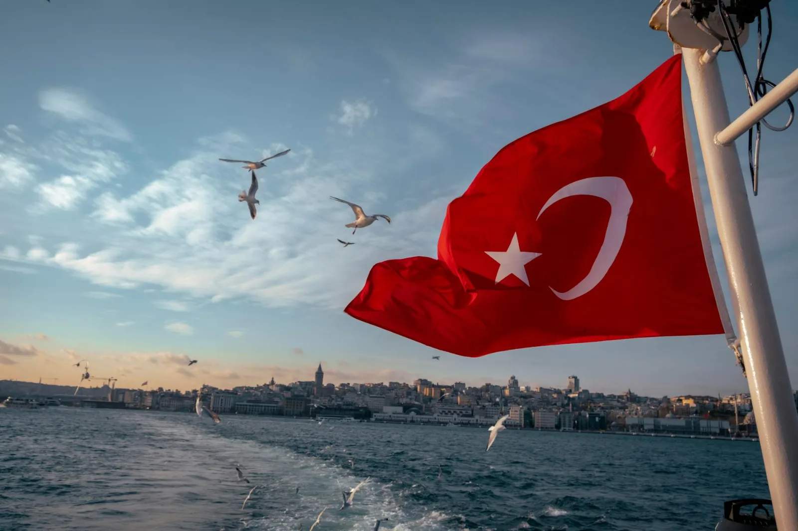 Turkisk flagga viftar från båt på Bosporen med Istanbul i bakgrunden och måsar i luften.